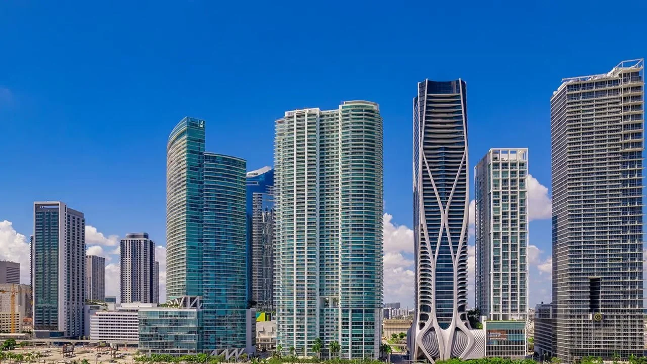 The tall white and glass 900 Biscayne Bay tower in Downtown Miami, beside the white curving exoskeleton of One Thousand Museum and other high-rises under a blue sky.
