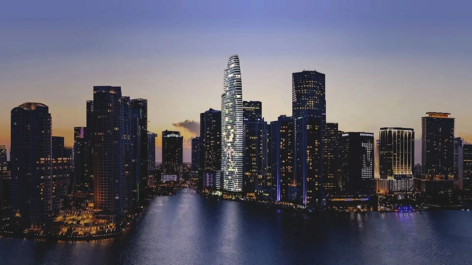 Rendering of the tapering Aston Martin Residences tower lit up at dusk among the Downtown Miami towers on the waterfront.
