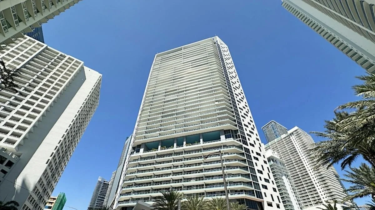The Brickell House condo tower seen from street level between neighbouring buildings and palm trees.