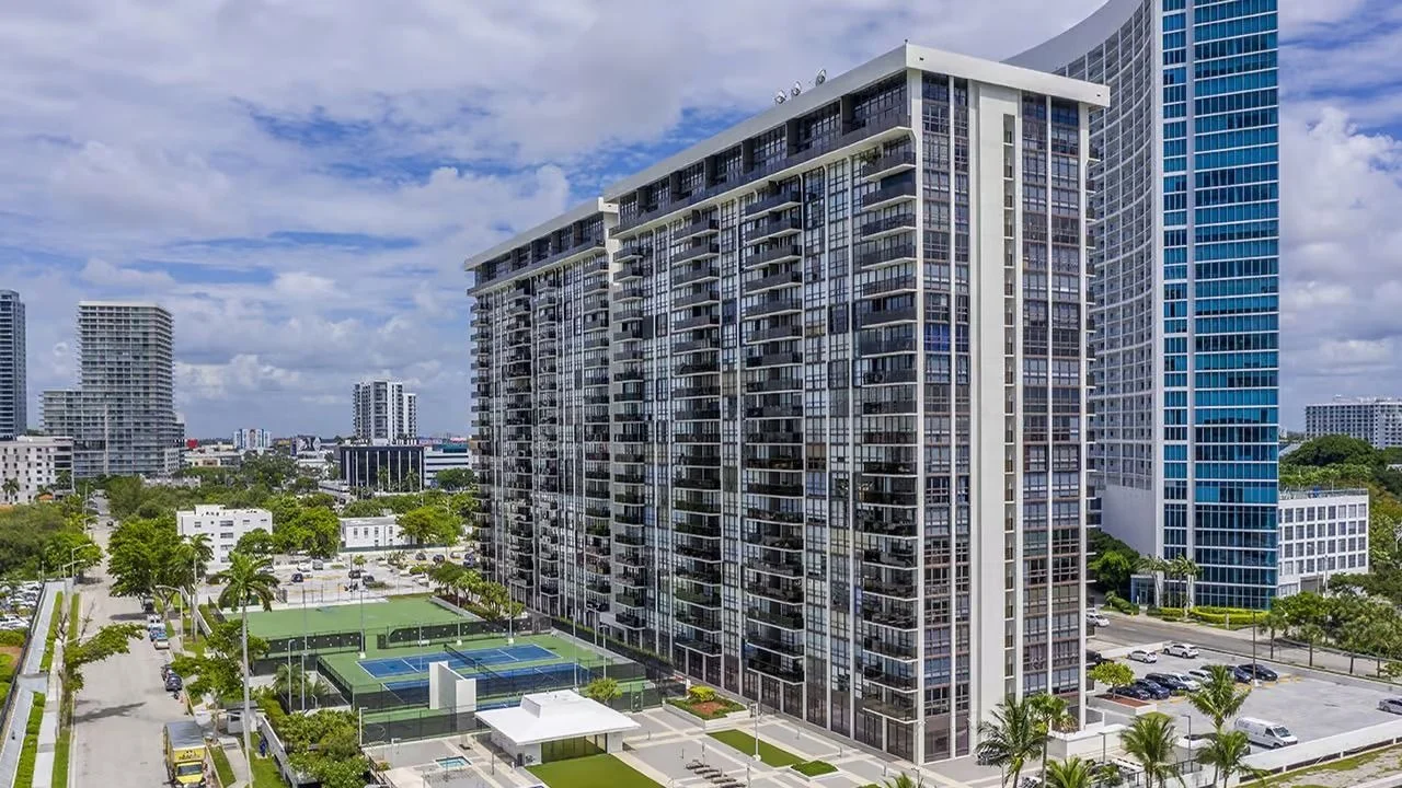 The long Charter Club condominium building in Edgewater with rows of balconies, seen from above beside tennis courts and a blue glass tower.