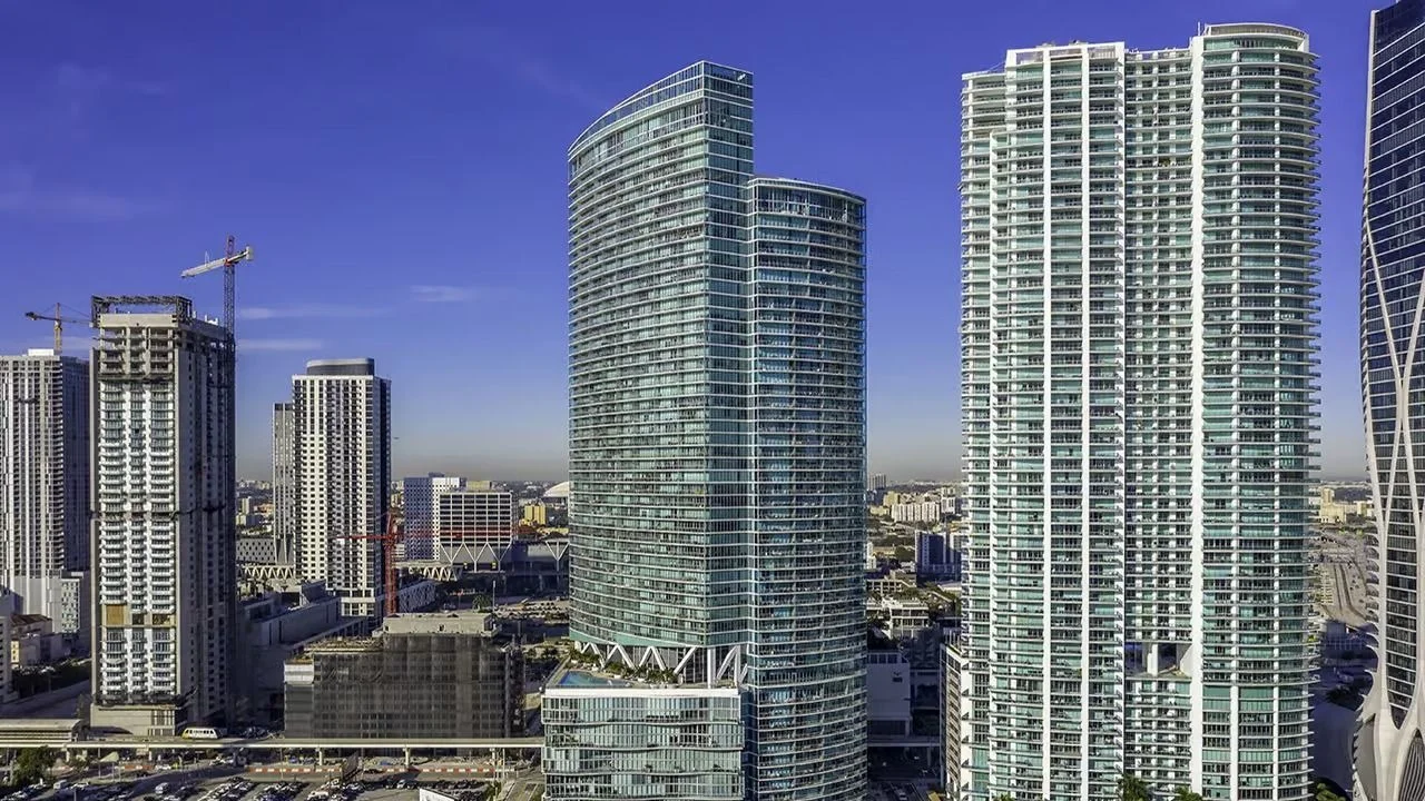 The curved blue glass Marina Blue tower in Downtown Miami, beside a taller white condo tower, with construction cranes on the left.