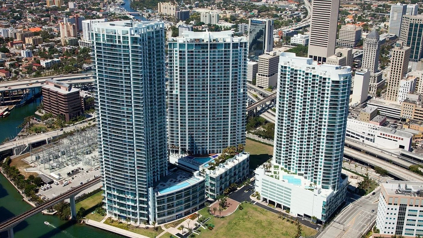 Aerial view of the Mint at Riverfront tower and its neighbouring towers beside the Miami River, with highway ramps and Downtown Miami around them.