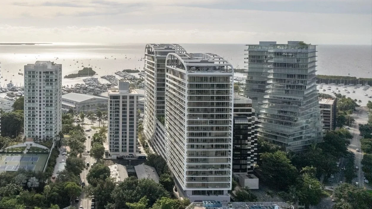 Coconut Grove from above, with the white Mr. C Residences towers with arched rooftops at the centre, the twisting Grove at Grand Bay towers to the right and Biscayne Bay behind.