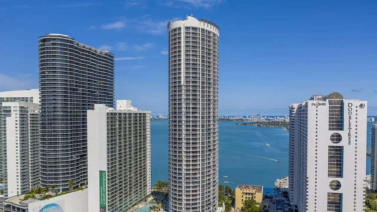 The tall, rounded white Opera Tower in Edgewater, between the curved Aria on the Bay tower and the DoubleTree hotel, with Biscayne Bay behind.