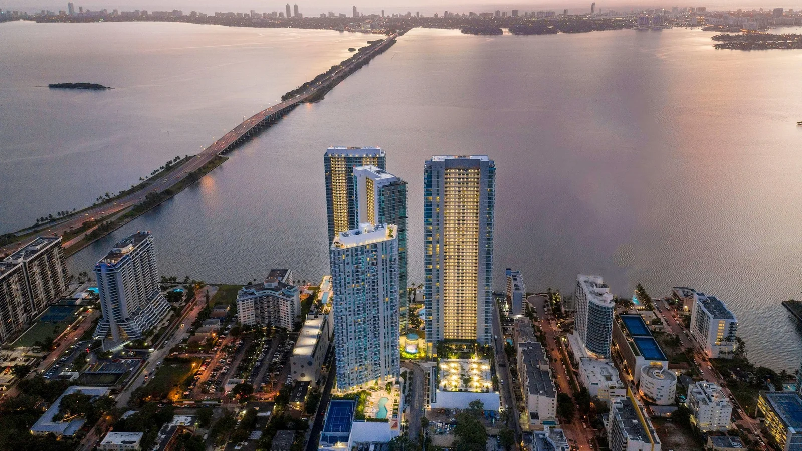 The Paraiso District towers in Edgewater, including Paraiso Bayviews, lit at dusk beside Biscayne Bay and a causeway.