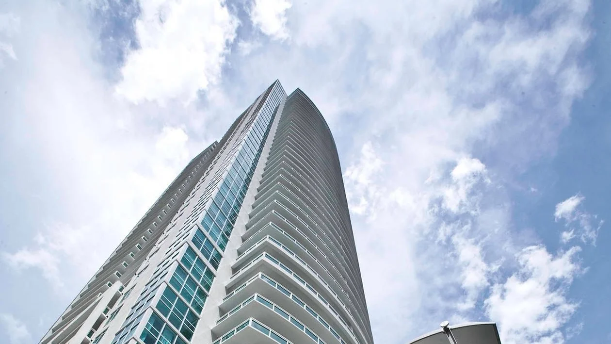 The tall curved facade of the Plaza on Brickell tower seen from below against white clouds.