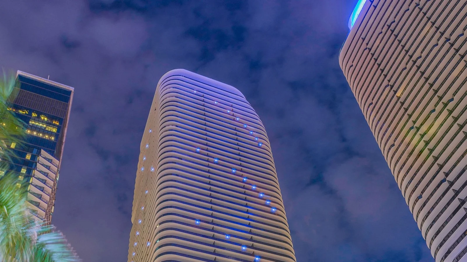 The rounded top of the SLS LUX Brickell tower lit at night, seen from street level between neighbouring towers.